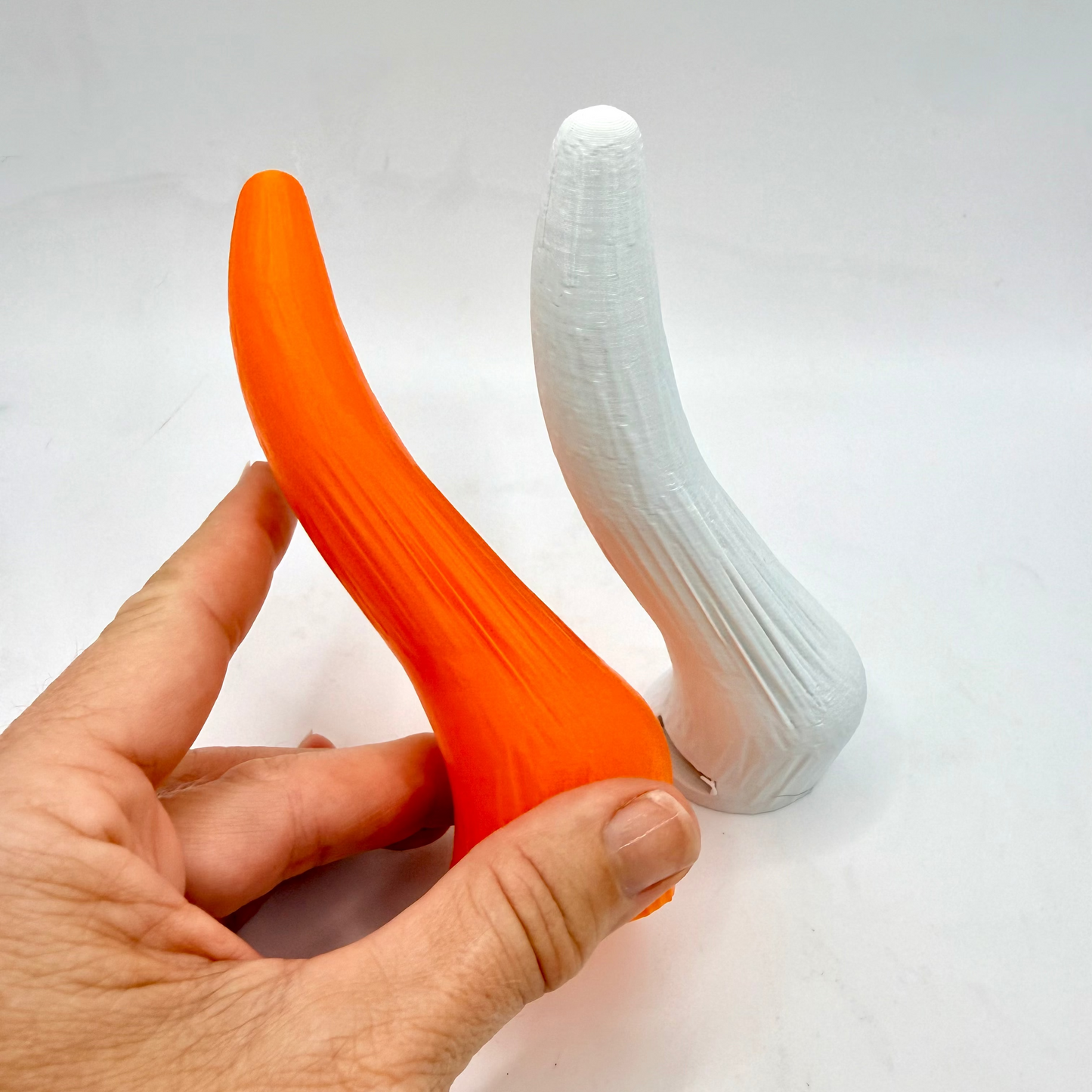 An adult novelty product shaped like a curved bone, shown in two colors: one held in a hand as bright orange, and one standing upright in white. The hand is visible on the left side of the frame against a plain white background.